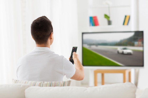 concepto de ocio, tecnología, deportes de motor, deporte y personas - hombre con control remoto viendo carreras de autos en la televisión en casa