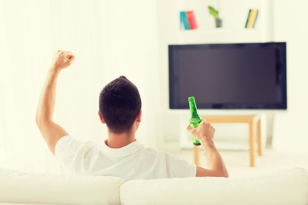 concepto de ocio, tecnología, medios de comunicación y personas: hombre viendo televisión, equipo de apoyo y bebiendo cerveza de botella en casa desde atrás