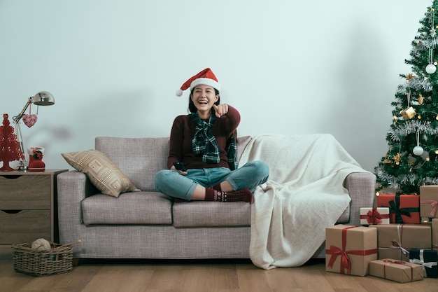 concepto de ocio de vacaciones de programa de navidad de televisión. mujer sonriente sentada en el sofá con control remoto de tv en el sofá de casa con fondo blanco de árbol de navidad. pantalla de punto de risa de niña alegre