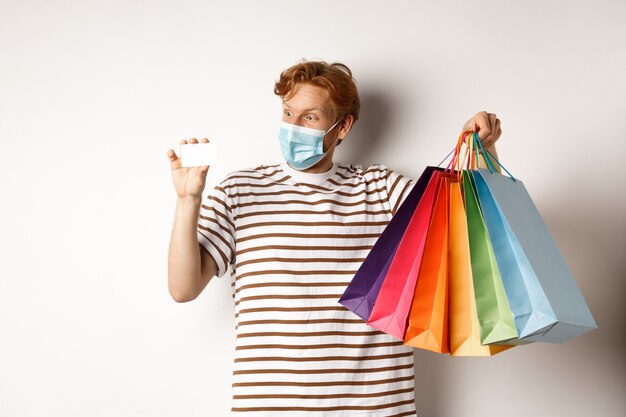 Concepto de oferta especial y pandemia. Hombre pelirrojo feliz en mascarilla mostrando bolsas de compras y tarjetas de crédito de plástico, de pie sobre fondo blanco.