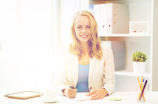 concepto de oficina, negocios, educación, tecnología e Internet - mujer de negocios sonriente o estudiante sentada en la mesa y escribiendo en un cuaderno