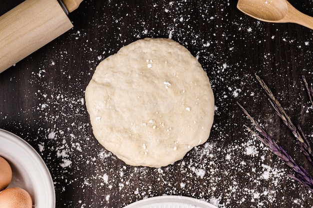 Concepto de pan una masa de bollo una pequeña espátula de madera un rodillo y un bol con unos cuantos huevos