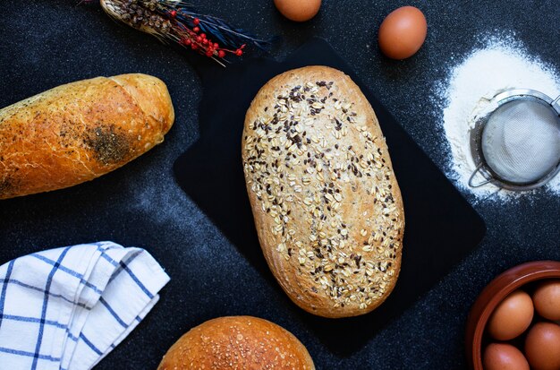 Concepto de panadería. Una variedad de diferentes tipos de pan sobre un fondo oscuro. Huevos, harina, espigas. Vista superior (endecha plana)