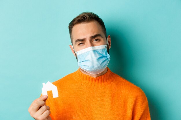 Concepto de pandemia de bienes raíces y coronavirus. Primer plano de un hombre triste en la mascarilla que muestra la mascarilla y haciendo muecas molesto, de pie