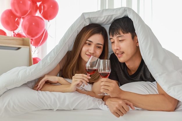 Concepto de pareja, amor y día de San Valentín. Retrato de una pareja en la cama con una manta blanca y sosteniendo un vaso de vino tinto en el dormitorio con globo rojo.