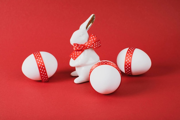 Concepto de Pascua Conejo de porcelana con tres huevos blancos decorados con cinta roja sobre fondo de papel rojo Copiar espacio