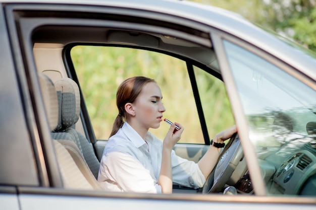 Concepto de peligro al conducir Mujer joven conductor adolescente pelirroja pintándose los labios haciendo aplicar maquillaje mientras conduce el coche