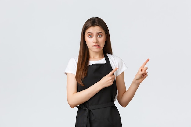 Concepto de pequeña empresa, empleados y cafetería. Barista lindo nervioso mordiendo el labio ansioso y apuntando la esquina superior derecha insegura. Vendedora novato indeciso preguntando sobre el producto