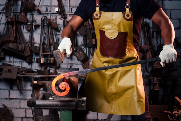 Concepto de pequeña empresa de la industria del martillo. El hombre afroamericano vestido con ropa histórica está martillando el yunque. Un herrero forja un producto de metal.