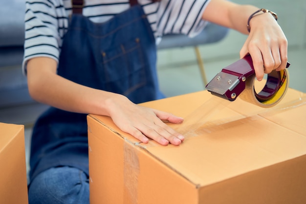 Concepto de pequeña empresa de inicio, joven mujer asiática propietaria trabajando y empacando en la caja al cliente en el sofá de la oficina en casa, el vendedor prepara la entrega.