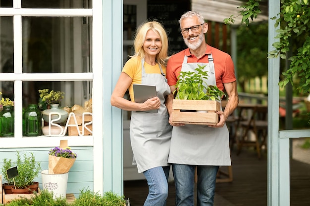 Concepto de pequeña empresa sonriente pareja de propietarios de invernaderos maduros posando cerca de su propia cafetería
