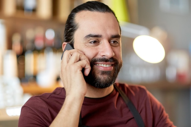 concepto de pequeñas empresas, personas y servicios - hombre feliz o camarero en delantal llamando al teléfono inteligente en el bar