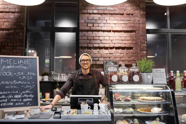 concepto de pequeñas empresas, personas y servicios - vendedor feliz o barman en el mostrador con caja en café vegano o cafetería