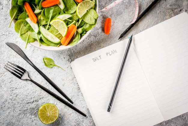 Concepto de pérdida de peso del plan de dieta, ensalada de verduras frescas con tenedor, cuchillo, bloc de notas, vista superior de espacio de copia de mesa de piedra gris