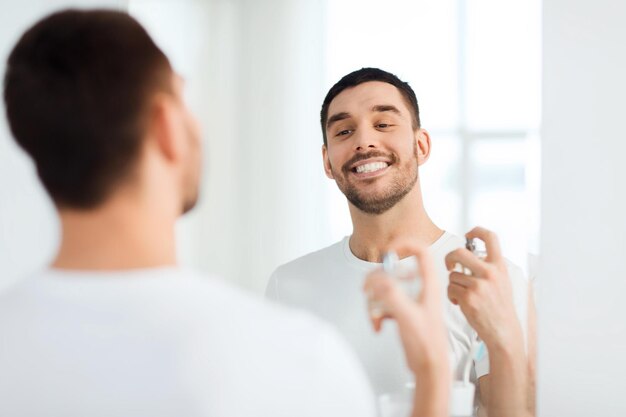 Foto concepto de perfumería, belleza y personas - joven sonriente feliz con perfume mirando al espejo usando aroma en el baño