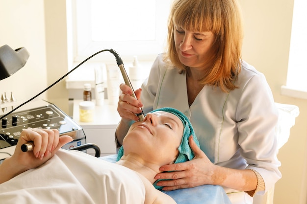 Concepto de personas, belleza, spa, cosmetología y tecnología: la cosmetóloga hace que el procedimiento sea una terapia de microcorriente de la piel facial de una hermosa y joven mujer en un salón de belleza