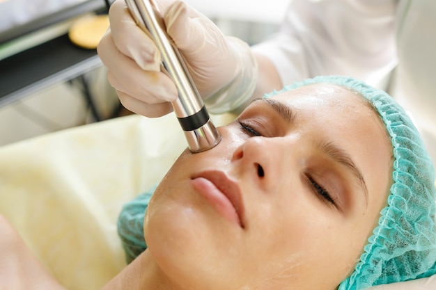 Concepto de personas, belleza, spa, cosmetología y tecnología: la cosmetóloga hace que el procedimiento sea una terapia de microcorriente de la piel facial de una hermosa y joven mujer en un salón de belleza