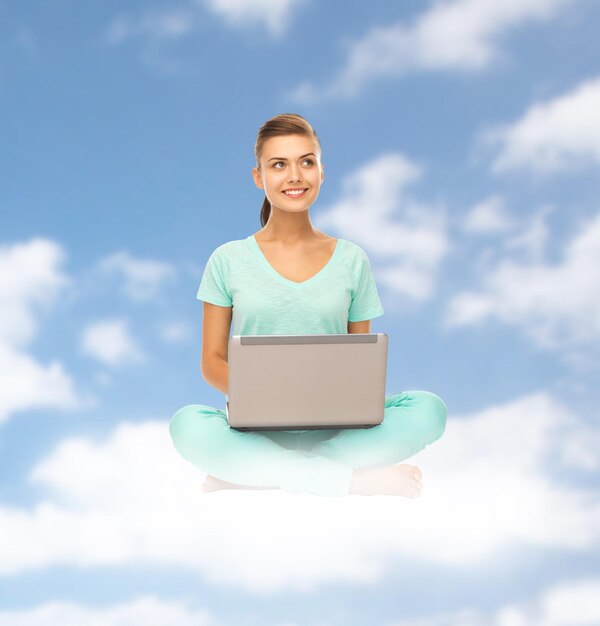 concepto de personas, computación y tecnología - mujer joven feliz con computadora portátil sentada en la nube sobre fondo de cielo azul
