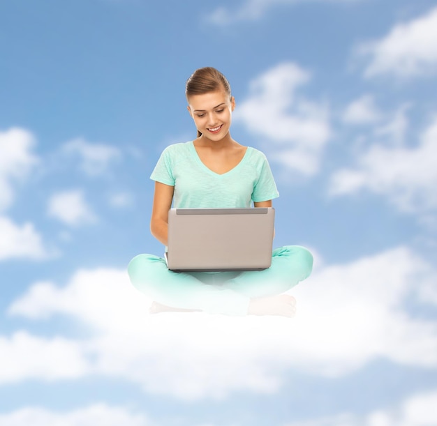 concepto de personas, computación y tecnología - mujer joven feliz con computadora portátil sentada en la nube sobre fondo de cielo azul