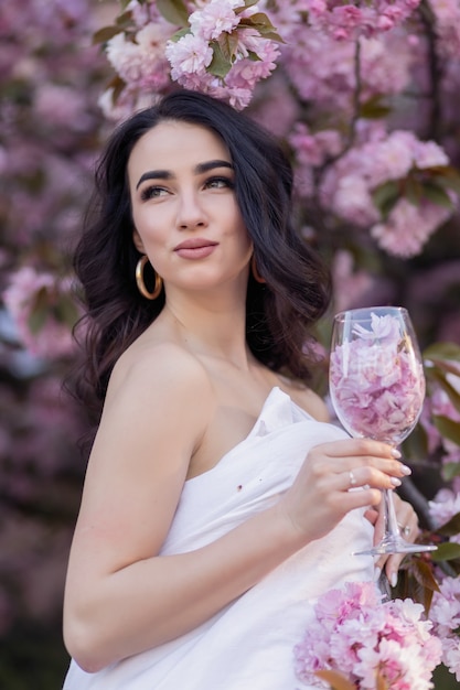 Concepto de personas, descanso, comodidad y ocio - Mujer joven por las calles de la tarde envuelta en una manta. Hermosa joven en el parque y un árbol en flor sakura sosteniendo un vaso con pétalos