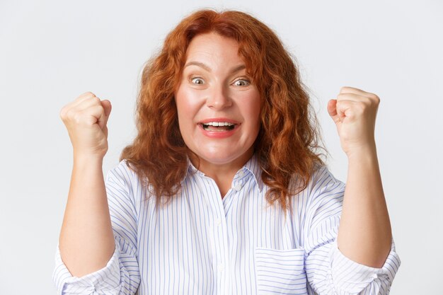 Concepto de personas, emociones y estilo de vida. Primer plano de la mujer pelirroja de mediana edad ganadora afortunada celebrando la victoria, mirando emocionada y emocional, bomba de puño de regocijo, pared blanca.