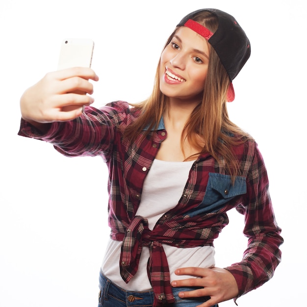 Concepto de personas, estilo de vida y tecnología: bonita adolescente con sombrero, tomando selfies con su teléfono inteligente, aislado en blanco