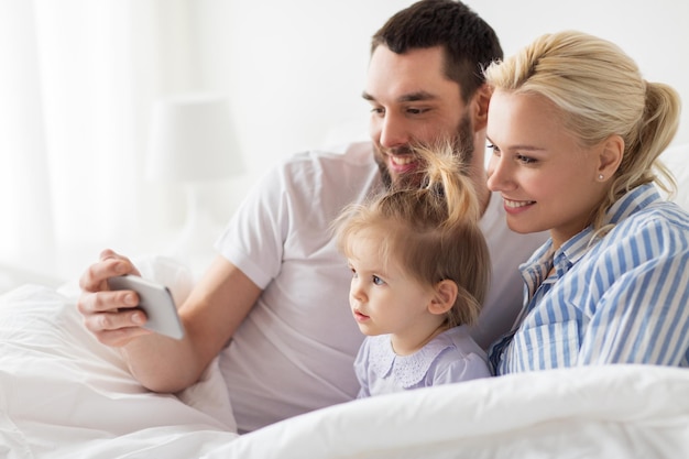 concepto de personas, familia y tecnología: madre feliz, padre y niña con teléfono inteligente en la cama en casa