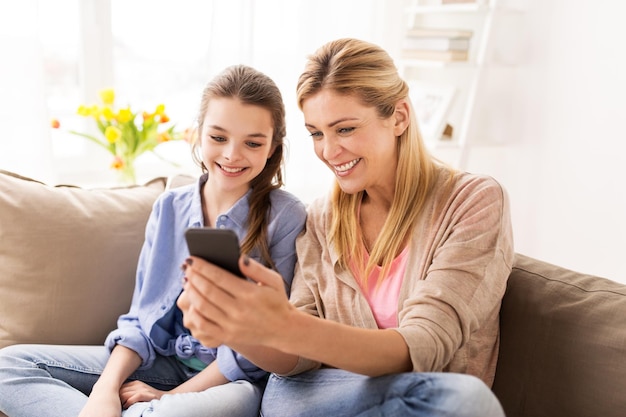 concepto de personas, familia y tecnología: niña feliz y madre con teléfono inteligente en casa