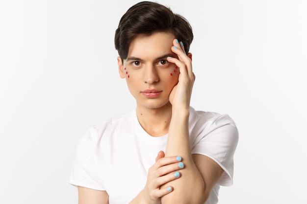 Concepto de personas, lgbtq y belleza. Primer plano de hermoso hombre queer tocando la cara con los dedos con esmalte de uñas azul, de pie sobre blanco.