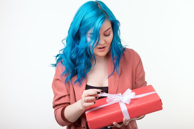 Concepto de personas, moda y vacaciones - hermosa chica con cabello azul de pie con caja de regalo en la pared blanca.