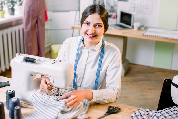 Concepto de personas, modista, sastre y moda: joven diseñadora de moda en su sala de exposición utilizando una máquina de coser