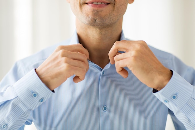 concepto de personas, negocios, moda y ropa - cerca de un hombre sonriente vistiéndose y ajustando el cuello de la camisa en casa