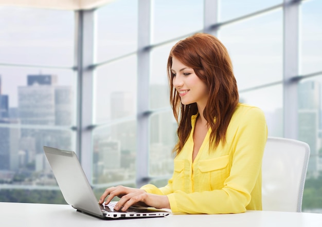 concepto de personas, negocios y tecnología - mujer joven sonriente con computadora portátil sentada en la mesa sobre el fondo de la ventana de la oficina