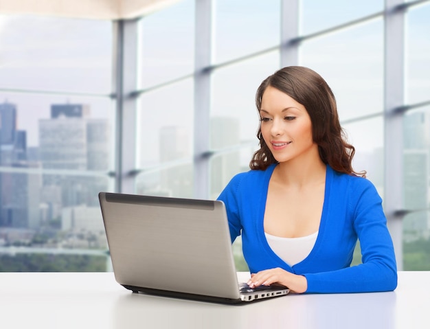 concepto de personas, negocios y tecnología - mujer sonriente vestida de azul con computadora portátil sentada en la mesa sobre el fondo de la ventana de la oficina