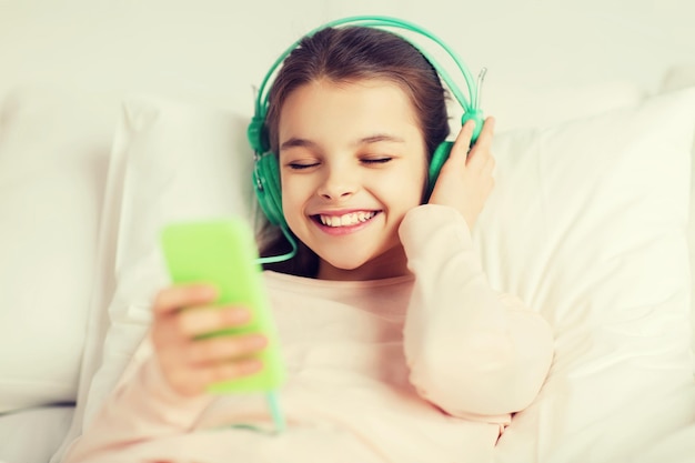 concepto de personas, niños, descanso y tecnología - feliz niña sonriente acostada despierta con smartphone y auriculares en la cama escuchando música en casa