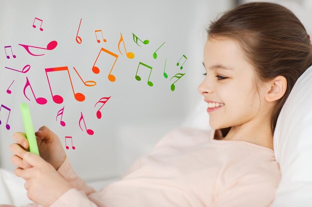 Foto concepto de personas, niños, música y tecnología - feliz niña sonriente acostada despierta con un smartphone en la cama en casa con notas musicales