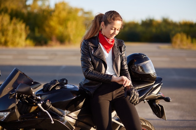 Concepto de personas, protección y conducción. Bonita mujer motociclista se pone guantes protectores, casco, se prepara para conducir en moto, posa contra la naturaleza borrosa