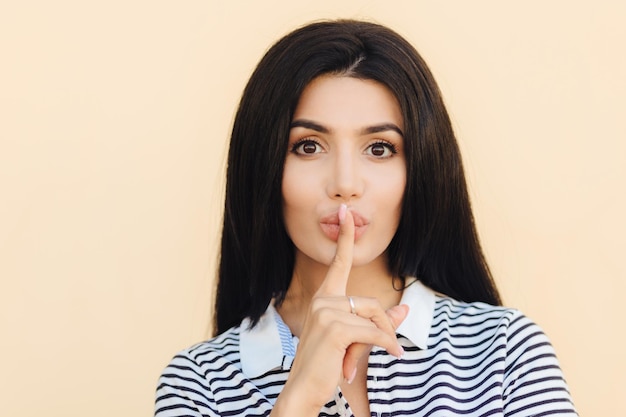 Concepto de personas y silencio Atractiva mujer con signo de silencio mantiene el dedo índice en los labios pide silencio mientras alguien duerme vestido con ropa informal gestos interiores Personas y conspiración