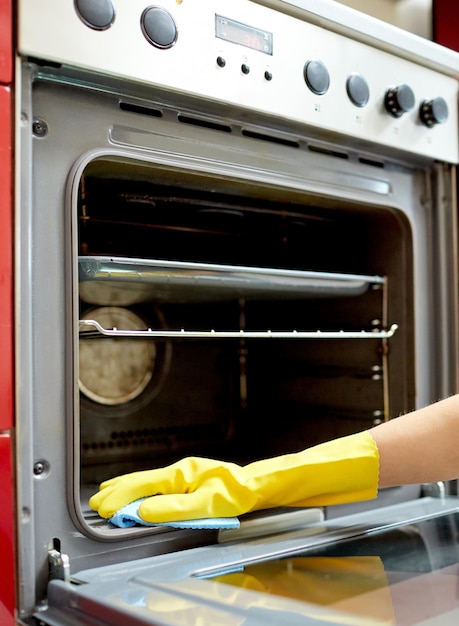 Concepto de personas, tareas domésticas y tareas domésticas: cerca de la mano de la mujer en un guante protector con un horno de limpieza de trapos en la cocina de su casa