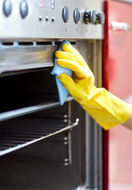 Concepto de personas, tareas domésticas y tareas domésticas: cerca de la mano de la mujer en un guante protector con un horno de limpieza de trapos en la cocina de su casa