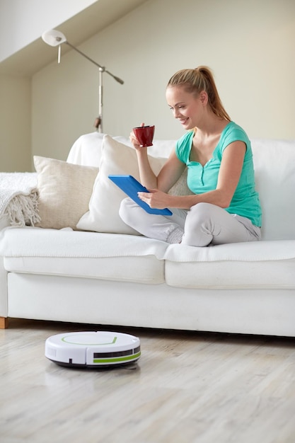 concepto de personas, tareas domésticas y tecnología - mujer feliz con computadora de tablet pc y aspiradora robot bebiendo té en casa