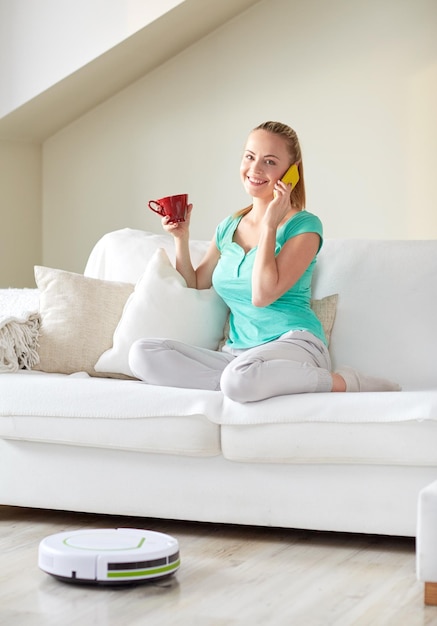 concepto de personas, tareas domésticas y tecnología - mujer feliz con smartphone y aspiradora robot bebiendo té en casa