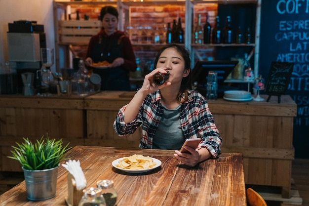 concepto de personas y tecnología. feliz mujer china asiática con smartphone bebiendo cerveza y leyendo mensajes en el bar. joven camarera en delantal trabajando en segundo plano preparando comida en el pub del mostrador