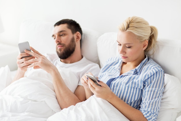 Concepto de personas, tecnología, Internet, relaciones y comunicación - pareja con teléfonos inteligentes en la cama en casa