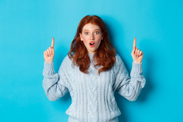 Concepto de personas y vacaciones de invierno. Adolescente pelirroja sorprendida apuntando con el dedo hacia arriba, mostrando publicidad y mirando impresionado a la cámara, fondo azul.