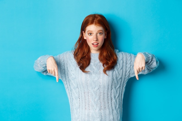 Concepto de personas y vacaciones de invierno. Linda adolescente señalando con el dedo hacia abajo, mostrando publicidad, de pie sobre fondo azul.