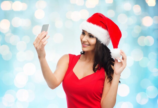 concepto de personas, vacaciones, navidad y tecnología - hermosa mujer sexy con sombrero rojo de santa tomando una foto selfie por teléfono inteligente sobre fondo de luces azules