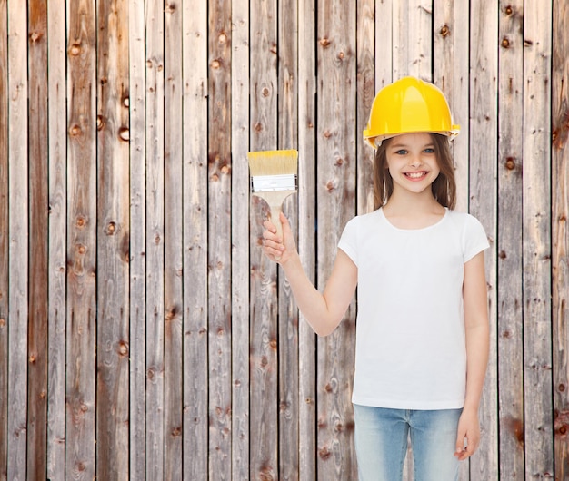 concepto de pintura, construcción, infancia y personas - niñita sonriente con casco protector con pincel sobre fondo de valla de madera