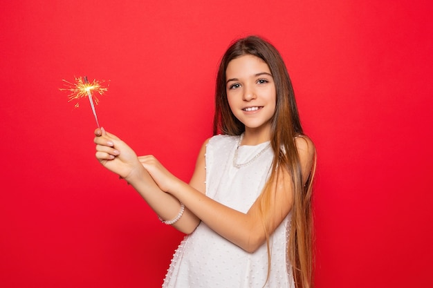 Concepto de pirotecnia y personas - sonriente joven o adolescente mujer feliz con luces de bengala celebran en vestido blanco sobre fondo rojo. mirando a la camara y sonriendo