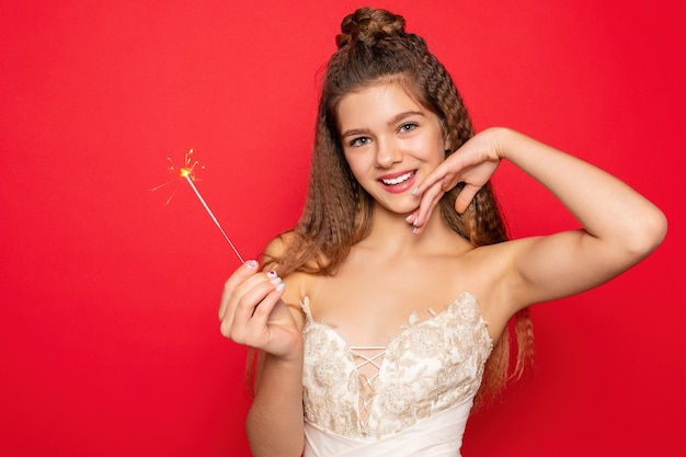 Concepto de pirotecnia y personas - sonriente joven o adolescente mujer feliz con luces de bengala celebran en vestido blanco sobre fondo rojo.